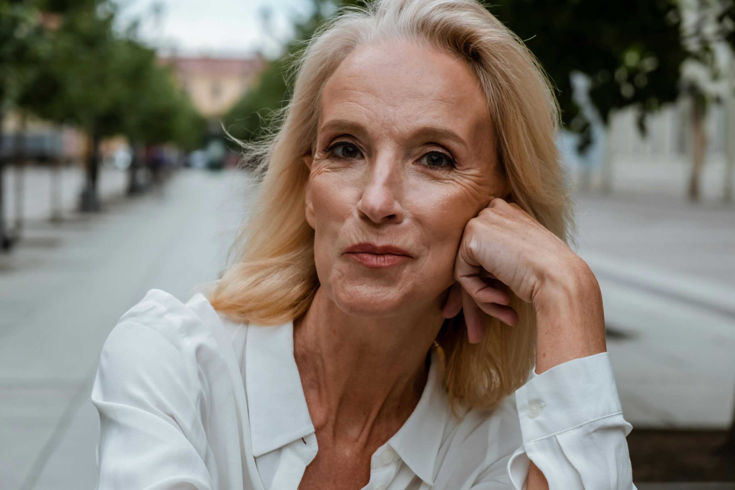 Portrait einer attraktiven älteren Frau. Sie lächelt in die Kamera und hat den Kopf seitlich auf die Hand aufgestützt. Im Hintergrund eine Allee.