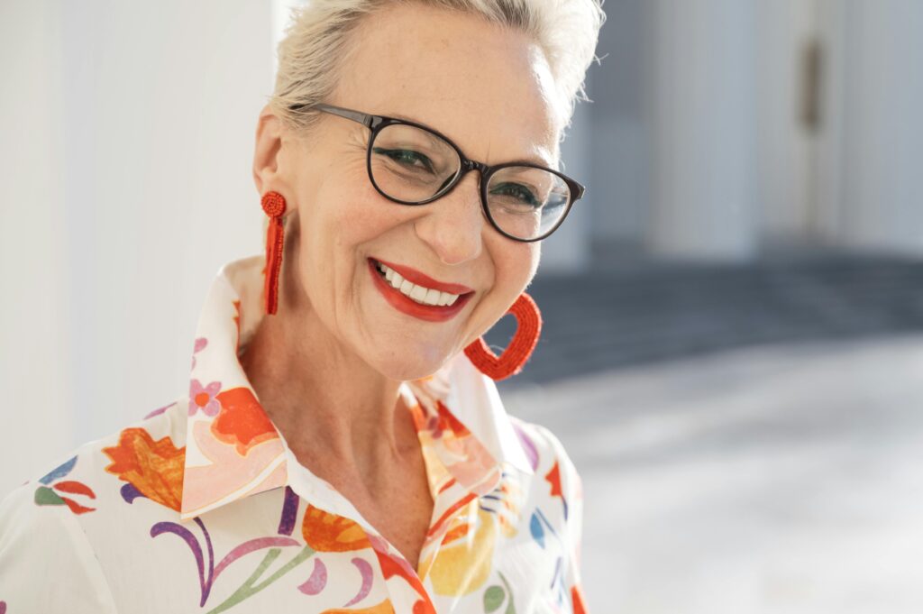 Portrait einer glücklich lächelnden attraktiven älteren Frau vor grauem, diffusem Hintergrund. Sie trägt große orangefarbene Ohrringe, ein bunt gemustertes Kleid und eine Brille. Beauty & Anti-Aging