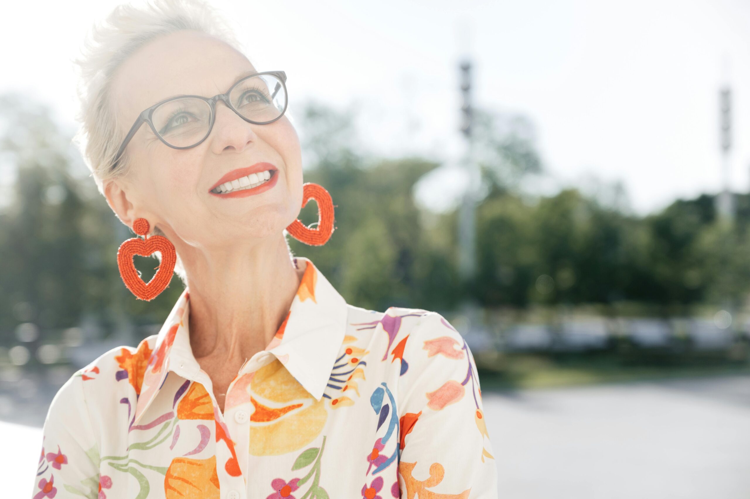 Portrait einer lächelnden, attraktiven älteren Frau mit frischem Teint. Sie trägt große, orangefarbene Ohrringe in Herzform, ein bunt gemustertes Kleid und eine Brille.. Sie schaut nach oben. Kollagen ab 50 - Beauty & Anti-Aging
